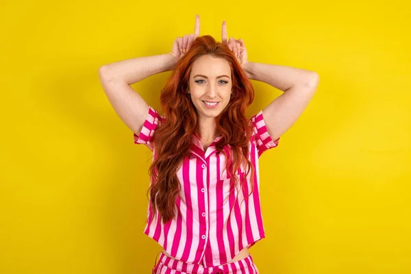 黄色いスタジオの背景にピンクのパジャマを着用している若い赤毛の女性は 頭のジェスチャーの指を示し 馬鹿げてかわいいポーズ — ストック写真