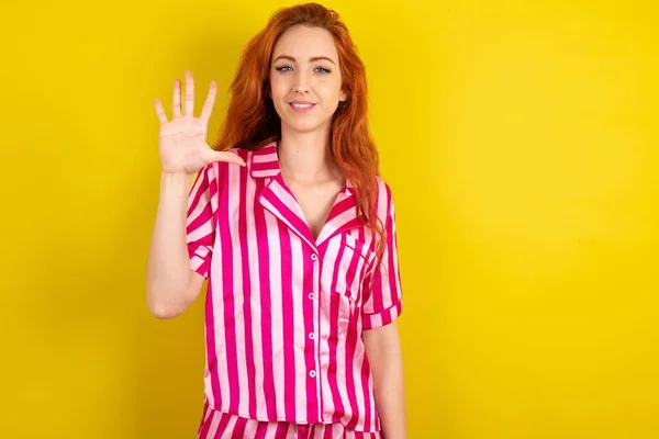 黄色いスタジオのバックグラウンドでピンクのピンクのパジャマを身に着けている若い赤毛の女性は自信と幸せに微笑みながら指の番号を5つ指で指摘します — ストック写真