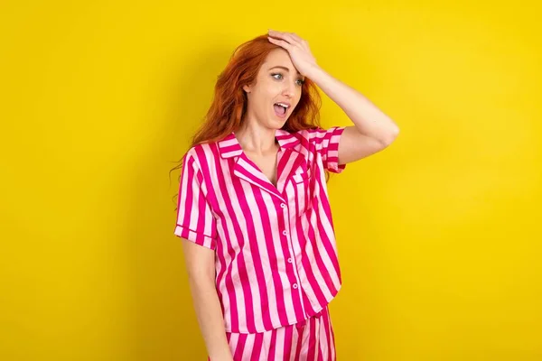 黄色いスタジオの背景にピンクのピンクのパジャマを着用した若い赤毛の女性は 間違いのために頭に驚いた 誤りを覚えています 忘れた 悪い記憶の概念 — ストック写真
