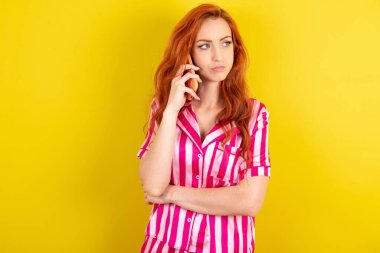 Sarı arka planda pembe pijama giyen, üzgün, genç, kızıl saçlı bir kadın akıllı telefondan konuşuyor. İletişim konsepti.