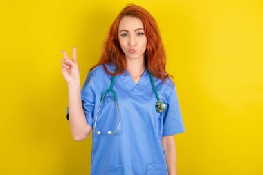 Sarı stüdyo arka planında genç kızıl saçlı doktor dudaklarını katlanmış V işaretleri ile barışçıl bir jest yapıyor. Vücut dili kavramı
