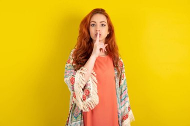 Sarı stüdyo arka planında duran kızıl saçlı kadın sessiz hareketler yapıyor, parmaklarını dudaklarının üzerinde tutuyor. Sessizlik ve gizli konsept.