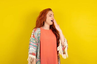 Sarı stüdyo arka planında duran kızıl saçlı kadın bütün gününü işte geçirdikten sonra yorgun ve esniyor..