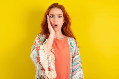 Şok olmuş kızıl saçlı bir kadın sarı stüdyo arka planında duruyor. Büyük bir şaşkınlıkla bakıyor. Beklenmedik haberler, yüz ifadeleri konseptiyle şaşkına dönüyor..