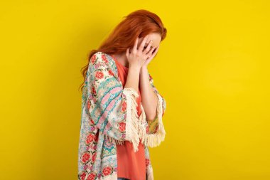 Kederli kızıl saçlı kadın sarı stüdyo arka planında duruyor ve elleriyle yüzünü kapatıyordu..