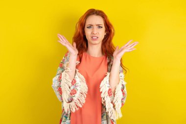 Sarı stüdyo arka planındaki kızgın kızıl saçlı kadın şaşkınlık içinde jestler yapıyor, tatminsizlikle kaşlarını çatıyor..