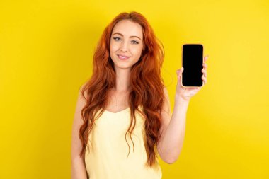 Sarı arka planda sarı tişört giyen kızıl saçlı güzel bir kadın. Boş ekran ile cep telefonunu tut