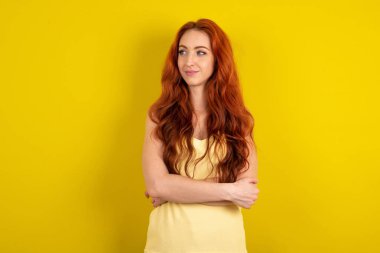 Sarı stüdyo arka planında sarı tişört giyen güzel, kızıl saçlı kadın ellerini göğsünün üzerinden uzatıyor. Mutlu bir şekilde yan tarafa bakıyor.