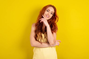 Sarı stüdyo arka planında sarı tişört giyen güzel kızıl saçlı kadın mutlu bir şekilde gülüyor, çenesinde elini tutuyor, olumlu duygular ifade ediyor, gülümsüyor, tasasız bir yüz ifadesi var.