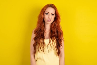 Sarı stüdyo arka planında sarı tişört giyen, memnun olmayan kızıl saçlı güzel bir kadının portresi sırıtıyor, dudakları çantasına bakıyor ve kameraya rahatsız edici bir şekilde bakıyor, hoş olmayan bir şey duyuyor.