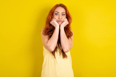 Sarı stüdyo arka planında sarı tişört giyen güzel kızıl saçlı bir kadın. Şaşırmış bir ifadeyle ellerini çenenin altında tutuyor. Dudaklarını katlıyor.