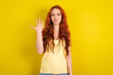 Sarı stüdyo arka planında sarı tişörtlü güzel kızıl saçlı kadın gülümsüyor ve arkadaş canlısı görünüyor, 5 ya da 5 numarayı gösteriyor, geriye doğru sayıyor.