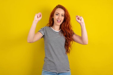 Şok olmuş kızıl saçlı kadın sarı arka planda çizgili tişört giyiyor. Şans piyangosunu kazanın ve el kaldırın ve bağırın.