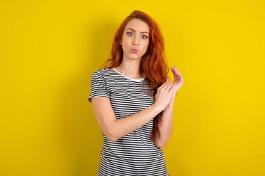 Sarı stüdyo arka planına çizgili tişört giyen kırmızı saçlı kadın avuçlarını ovuşturuyor ve kameraya inançsızlıkla bakıyor.