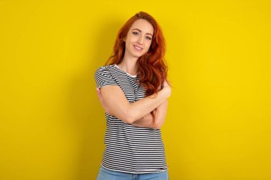 Sarı arka plan üzerinde çizgili gömlek giyen kızıl saçlı çekici bir kadın kendi vücudunu kucaklıyor, rahat pozlar veriyor. Şefkat ve kendine saygı kavramı