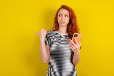 Sarı stüdyo arka planında çizgili gömlek giyen kızıl saçlı kadın başparmağıyla işaret ediyor ve boş alan gösteriyor, cep telefonunu mesaj göndermek için tutuyor..