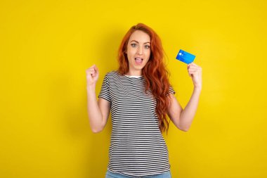 Şanslı kızıl saçlı kadının sarı stüdyo arka planına çizgili gömlek giyip kredi kartı tutan şanslı fotoğrafı. Ünlü.