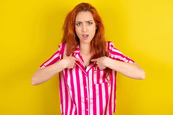 黄色いスタジオの背景にピンクのピンクのパジャマを着用した恥ずかしい赤毛の女性は 競争に参加するために選ばれることにショックを受け 何かについて躊躇して自分自身を示しています — ストック写真