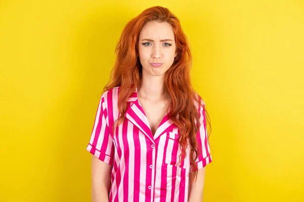 黄色いスタジオの背景にピンクのピンクのパジャマを身に着けている熱烈な陰鬱な拒絶された赤い髪の女性は問題と困難を抱え より低い唇を曲げ 絶望の中で目を閉じ うつ状態にある — ストック写真