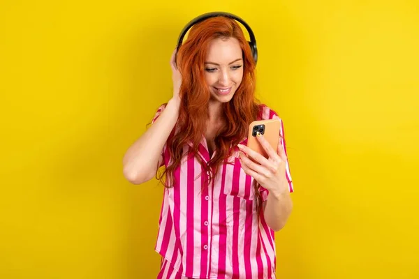 黄色いスタジオの背景にピンクのピンクのパジャマを着用したハッピー赤毛の女性は スマートフォンの画面に焦点を当てながら良い感じです ライフスタイル — ストック写真