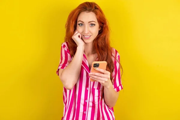 黄色いスタジオの背景にピンクのピンクのパジャマを身に着けているヨーヨーヨーの赤毛の女性は携帯電話装置が付いているポーズを 現代スマートフォンのテキスト メッセージをタイプし 自由時間の間に面白いビデオを見ます よいインターネット接続を楽しんで下さい — ストック写真