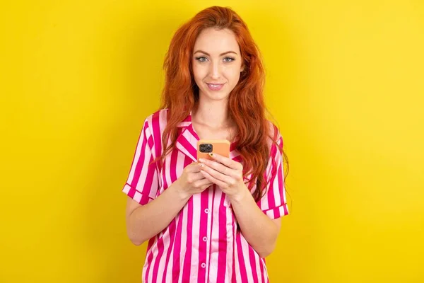 黄色いスタジオの背景にピンクのピンクのパジャマを身に着けている赤毛の女性は遠いコミュニケーションを楽しみ 携帯電話を使用し サーフの速い無制限のインターネット 楽しい笑顔を持っています オンラインで買い物をします — ストック写真
