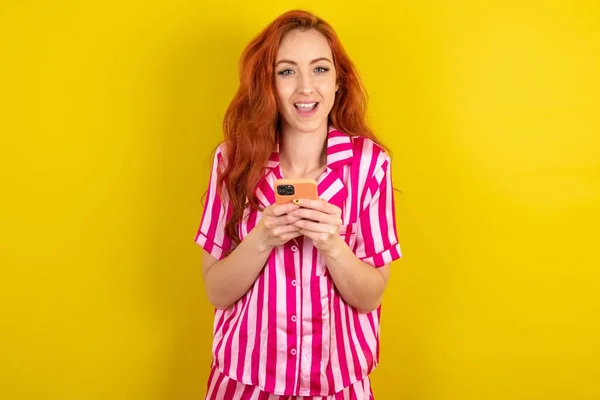 エキサイティングな赤毛の女性は スマートフォンを保持している黄色いスタジオの背景にピンクのパジャマを着て 良いニュースを受け取った後 カメラに驚きました — ストック写真