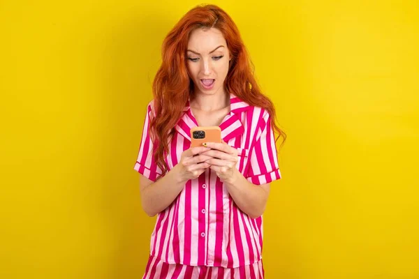 在黄色工作室背景下穿着粉色睡衣的红头发女人使用手机聊天 — 图库照片