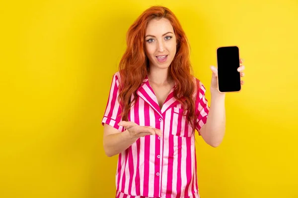 黄色いスタジオの背景にピンクのピンクのパジャマを着た若い赤毛の女性は 携帯電話を提示するスマートフォンで 広告コンセプト — ストック写真