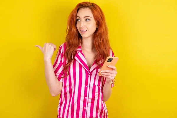 黄色いスタジオの背景にピンクのピンクのパジャマを身に着けている若い赤毛の女性は スマートフォンの指でテキストし 幸せな顔の笑顔で親指で示しています — ストック写真