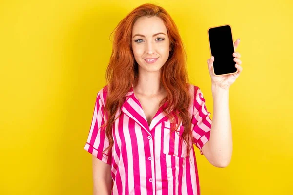 白い空のスクリーンが付いている携帯電話を握るとき黄色いスタジオの背景にピンクのパジャマを身に着けている若い赤毛の女性 — ストック写真