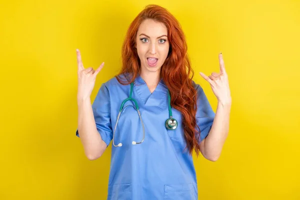 年轻红头发的医生女士在黄色工作室的背景下 使摇滚标志看起来自信而快乐 在派对上享受凉爽的音乐 身体语言概念 — 图库照片
