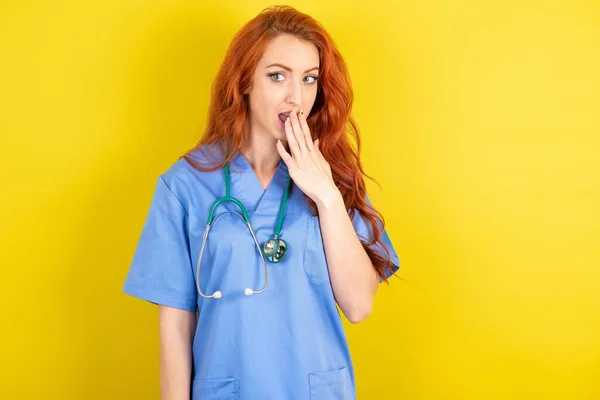 黄色いスタジオの背景にある若い赤毛の医師の女性は口を覆い カメラで不思議に見ます 予期しない噂を信じることができません — ストック写真
