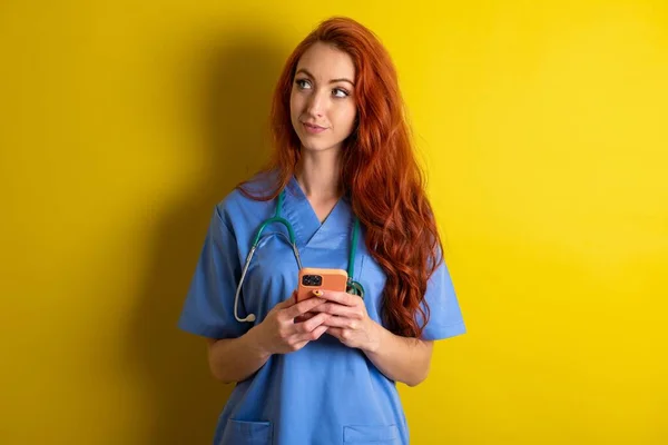 Ung Rödhårig Läkare Kvinna Över Gul Studio Bakgrund Innehar Mobiltelefon — Stockfoto