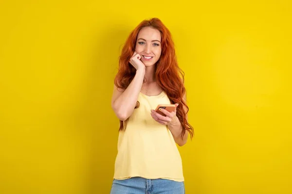 黄色いスタジオの背景に黄色いシャツを着ているヨーヨーヨーの赤毛の女性は携帯電話装置が付いているポーズを 現代スマートフォンのテキスト メッセージをタイプし 自由な時間の間に面白いビデオを見ます よいインターネット接続を楽しんで下さい — ストック写真