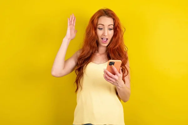 美しい赤毛の女性は 手の細胞の読書ニュースを保持している黄色いスタジオの背景の上に黄色のシャツを着ています — ストック写真