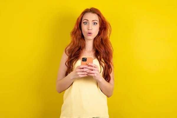 黄色いスタジオの背景に黄色いシャツを着ているショックを受けた赤毛の女性は 信じられないほどの大きなショッピング価格を宣伝する携帯電話の読書を開きます — ストック写真