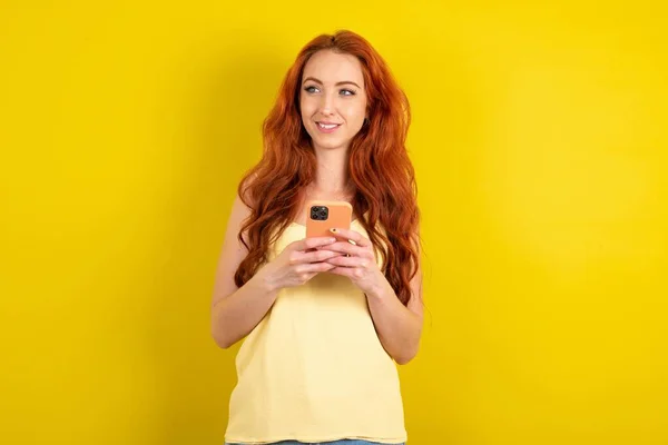 黄色いスタジオの背景に黄色いシャツを着ている美しい赤毛の女性は電話の手を読む良い若者のニュースは空のスペースの広告を見ます — ストック写真