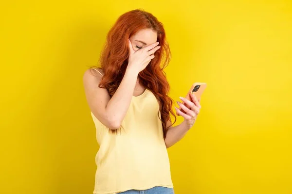 美しい赤毛の女性は黄色いスタジオの背景に黄色いシャツを着て スマートフォンを見て悲しい顔を抱えています — ストック写真
