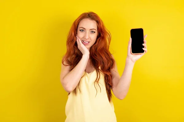 手で持っている黄色いスタジオの背景に黄色いシャツを着用する美しい赤毛の女性 現代技術を使用して感銘を受けた叫びを驚かせた手のひら 信じられないほどの予想外 — ストック写真
