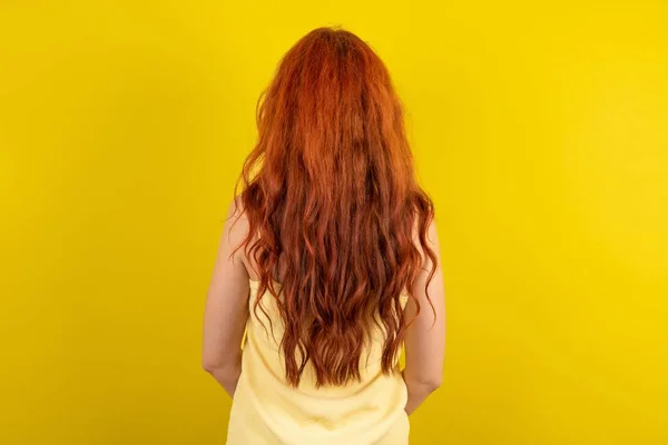 The back view of a beautiful red haired woman wearing yellow shirt over yellow studio background. Studio Shoot.