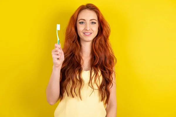 黄色いスタジオの背景に黄色いシャツを着た美しい赤毛の女性は 歯ブラシと微笑みを抱えています 歯科医療コンセプト — ストック写真