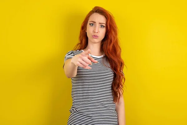 ショックを受けた赤毛の女性は イエロースタジオのバックグラウンドポイント上のストライプシャツを着て 驚くべき表情であなたに — ストック写真