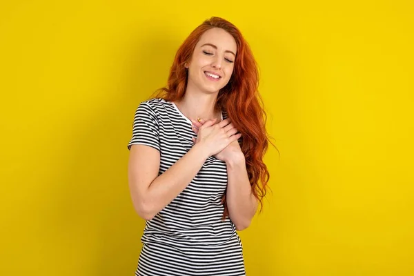 黄色いスタジオの背景にストライプのシャツを着ている赤毛の女性は幸せを表現し 喜んで笑い 心に手を保ちます — ストック写真