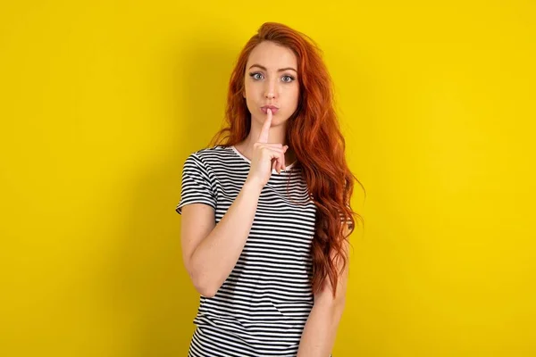 黄色いスタジオの背景にストライプのシャツを着用した驚異的な赤毛の女性は沈黙のジェスチャーを作り 唇の上に指を保ち カメラで神秘的に見えます — ストック写真