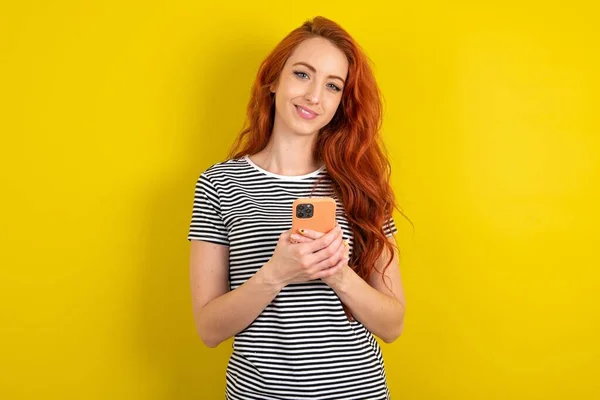 Рыжая Женщина Полосатой Рубашке Фоне Жёлтого Студийного Фона Использование Мобильного — стоковое фото