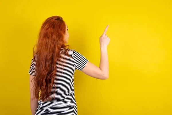 コピースペース リアビューにオブジェクトを指す黄色のスタジオの背景にストライプのシャツを着用した赤毛の女性 背中を向ける — ストック写真