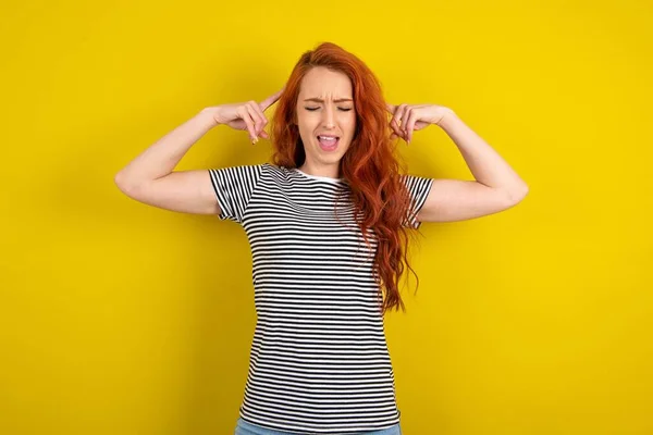 Photo of crazy red haired woman wearing striped shirt over yellow studio background screaming and pointing with fingers at hair closed eyes