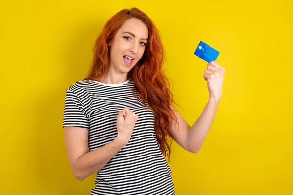 Excited happy positive cheerful smiling red haired woman wearing striped shirt over yellow studio background hold credit card raise fist in victory