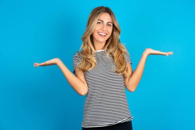 Neşeli, neşeli, iyimser, güzel bir kadın mavi arka plan üzerinde çizgili tişört giyiyor, elinde iki avuç içi fotokopi alanı tutuyor.